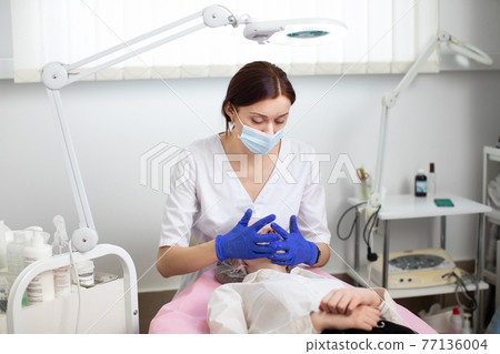Young female doctor cosmetologist in protective mask and blue rubber gloves makes facial massage and skin treatment for young woman client. Spa face massage in beauty salon, facial beauty treatment. 77136004