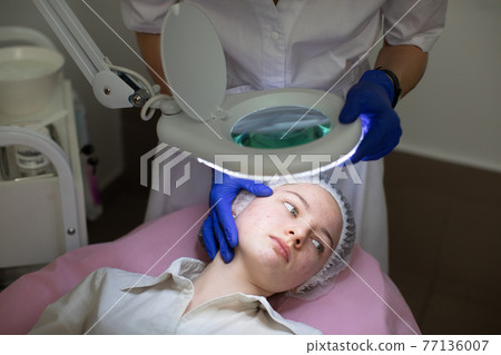 Close up of face of young teen girl with problem skin and acne, lying on the massage table in modern cosmetology clinic. Hands of female doctor beautician in gloves examines the skin of her patient. 77136007