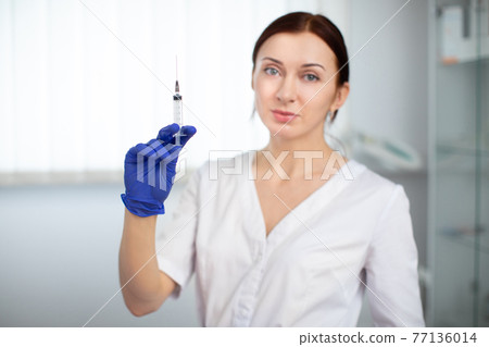 Young attractive doctor beautician with syringe. Close up of female cosmetologist indoors in medical parlor, holding syringe with skin care solution. 77136014