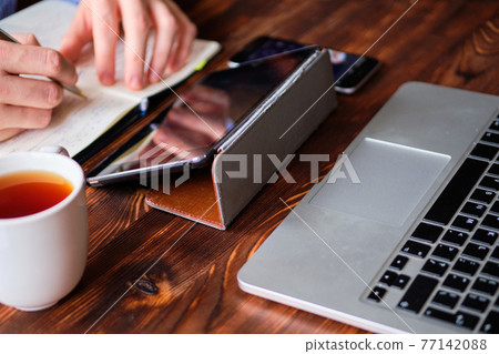 A man in a shirt makes notes in a diary and works on a laptop and tablet. Analytics activity concept. 77142088