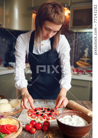 Woman in white shirt and apron putting the ingredients on the pizza. Pizza concept. Production and delivery of food Woman in white shirt and apron putting the ingredients on the pizza. Pizza concept. Production and delivery of food 77143282