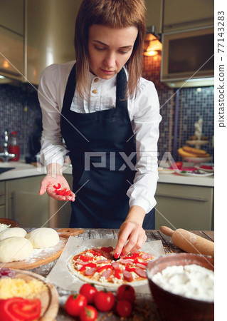 Woman in white shirt and apron putting the ingredients on the pizza. Pizza concept. Production and delivery of food Woman in white shirt and apron putting the ingredients on the pizza. Pizza concept. Production and delivery of food 77143283