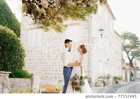 The bride and groom stand hugging under the pine branches near a beautiful brick house in the old town of Perast The bride and groom stand hugging under the pine branches near a beautiful brick house in the old town of Perast 77152495
