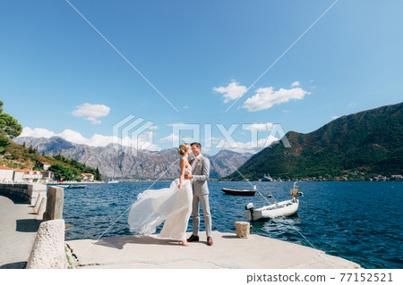 The bride and groom stand hugging on the pier in the Bay of Kotor, boats are next to them 77152521