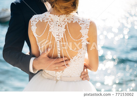 The bride and groom are standing and hugging on the pier near the sea, hand of the groom on bride's back, close-up 77153002