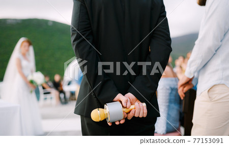 The host at a wedding party stands and holds a microphone behind his back, close-up  77153091