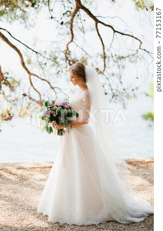 A sophisticated bride stands with a wedding bouquet in her hands under the branches of a tree by the sea  77153167