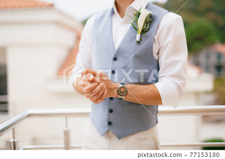 The groom in a shirt and vest with a boutonniere stands on the balcony in the morning before the wedding ceremony 77153180