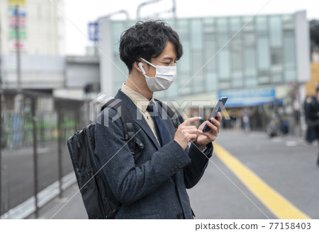 A man in his early thirties wearing headphones and wearing a mask to operate his smartphone 77158403