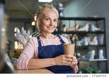 Portrait of happy mature woman in blue apron smiling at camera, drinking coffee or tea from disposable cup in her pottery studio 77164755