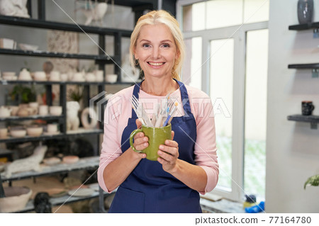 Ready to start. Portrait of happy woman in blue apron holding tools, brushes for creating handmade clay ceramic bowl in pottery workshop studio 77164780