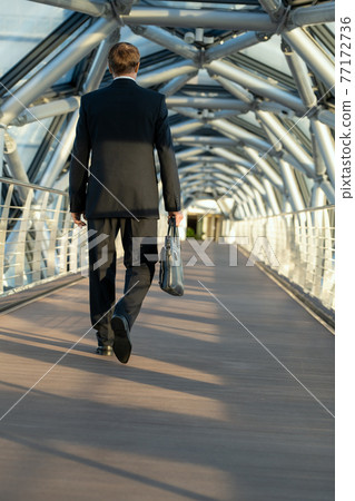 A man with black leather handbag walking down a long walkway 77172736