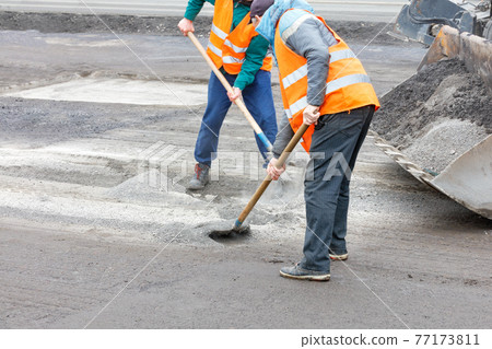 Two road workers rake old broken asphalt off the road and load it into a grader bucket. Two road workers rake old broken asphalt off the road and load it into a grader bucket. 77173811