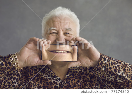 Funny senior man covering face with mobile phone with magnified picture of perfect smile Funny senior man covering face with mobile phone with magnified picture of perfect smile 77174840
