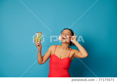 Happy brunette with closed eyes holding a summer cocktail and enjoying listening to music with headphones 77175673