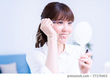 A woman applying mascara while looking at a hand mirror 77176903
