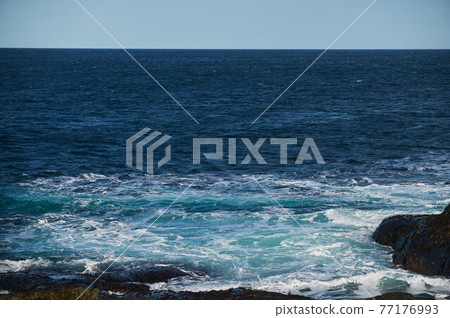 Small sea waves break against the stones of the shore. A bright sunny day and white foam of the waves. Melting glaciers as an environmental impact of air pollution. 77176993