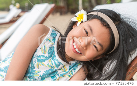 Portrait of Asian child girl leaning on sunbed on a beach of Thailand, black long hair with white flower at ear as decoration, smiling face, eyes looking at camera. Portrait of Asian child girl leaning on sunbed on a beach of Thailand, black long hair with white flower at ear as decoration, smiling face, eyes looking at camera. 77183476