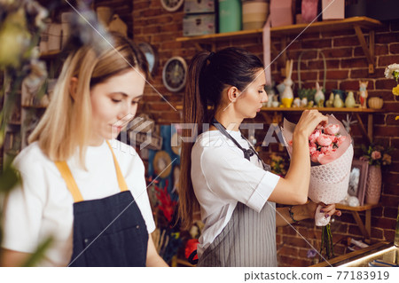 Woman florist together with assistant collect bouquets in flower shop. Woman florist together with assistant collect bouquets in flower shop. 77183919