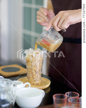 Professional barista preparing coffee on counter. Pouring coffee by hand from a measuring cup in a plastic cup (Closeup ). 77187995