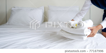 Hotel maid putting set of towel with white flower on the bed. Staff in blue uniform preparing room for hotel guest. 77187996