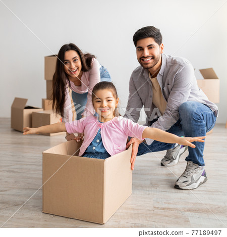 Real estate concept. Joyful eastern family having fun and celebrating moving day, parents riding daugher in box 77189497