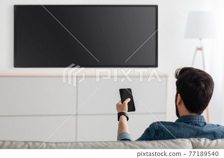 Young man watching television and using smart TV remote control application on smartphone, back view Young man watching television and using smart TV remote control application on smartphone, back view 77189540