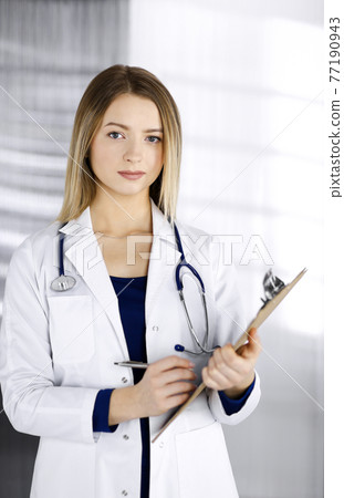 Young intelligent woman-doctor is prescribing some medicine for a patient, using a clipboard, while standing in the cabinet in a clinic. Female physician with a stethoscope at her working place Young intelligent woman-doctor is prescribing some medicine for a patient, using a clipboard, while standing in the cabinet in a clinic. Female physician with a stethoscope at her working place 77190943