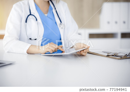 Unknown woman-doctor sitting in sunny clinic and using tablet pc, close-up. Data and best service in medicine Unknown woman-doctor sitting in sunny clinic and using tablet pc, close-up. Data and best service in medicine 77194941