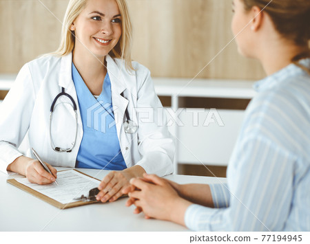 Woman-doctor and patient sitting and talking in hospital or clinic. Blonde therapist is cheerfully smiling. Medicine concept Woman-doctor and patient sitting and talking in hospital or clinic. Blonde therapist is cheerfully smiling. Medicine concept 77194945