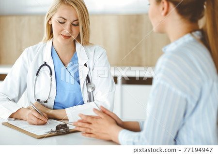 Woman-doctor and patient sitting and talking in hospital or clinic. Blonde therapist is cheerfully smiling. Medicine concept Woman-doctor and patient sitting and talking in hospital or clinic. Blonde therapist is cheerfully smiling. Medicine concept 77194947