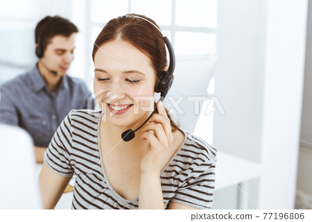Casual dressed young woman using headset and computer while talking with customers online. Group of operators at work. Call center, business concept 77196806