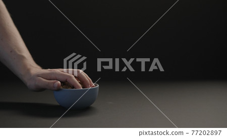 man take roasted salted pistachios from blue bowl on black paper background 77202897