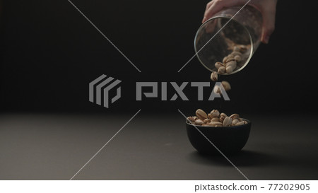 man pour roasted salted pistachios into black bowl on black paper background 77202905
