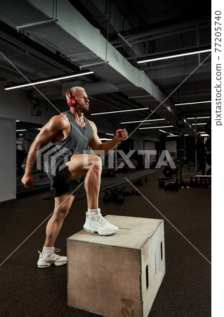 Sexy muscular men using the platform for his legs on a dark colorful background of gym. Sexy muscular men using the platform for his legs on a dark colorful background of gym. 77205740