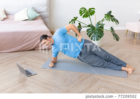 A middle-aged man trains at home online. He performs a side-bar exercise while looking at the laptop monitor. 77209502