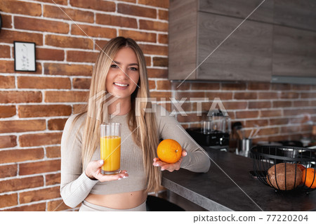Pretty girl holding glass of orange juice and orange fruit in hand at kitchen 77220204