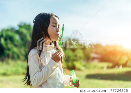 Portrait of funny lovely little Asian girl blowing soap bubbles in the park. 77224826
