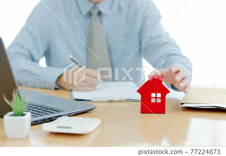 A House on desk envelope with man signing purchase documents in background. while hand complete the insurance policy, rental documents and loan document. concept guy buying new house A House on desk envelope with man signing purchase documents in background. while hand complete the insurance policy, rental documents and loan document. concept guy buying new house 77224873