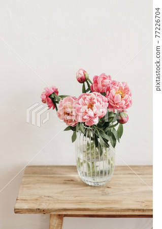 Floral still life scene. Pink peonies flowers, bouquet in glass vase on wooden table. White wall. Selective focus, blurred background. Wedding, birthday celeberation concept. Lifestyle vertical photo Floral still life scene. Pink peonies flowers, bouquet in glass vase on wooden table. White wall. Selective focus, blurred background. Wedding, birthday celeberation concept. Lifestyle vertical photo 77226704