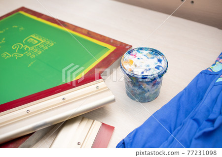 Serigraphy silk screen print process at clothes factory. Frame template stencil, squeegee and plastisol paint at table with cloth Serigraphy silk screen print process at clothes factory. Frame template stencil, squeegee and plastisol paint at table with cloth 77231098