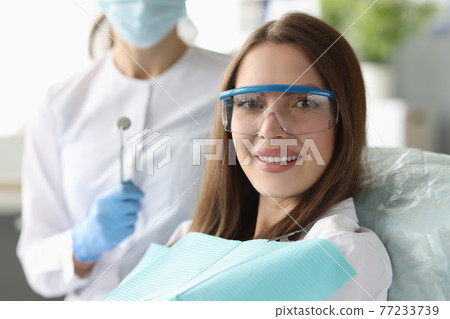 Woman patient in safety glasses sitting in dental chair near doctor Woman patient in safety glasses sitting in dental chair near doctor 77233739