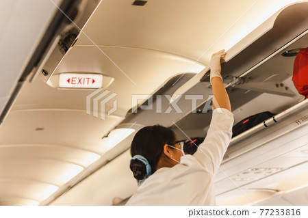 Flight attendant in protective masks and glove closing overhead compartment. Flight attendant in protective masks and glove closing overhead compartment. 77233816