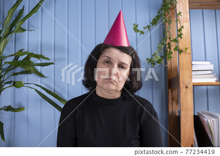 Sad Middle aged woman whit bitrthday hat and looking with sad eyes on camera. Senior woman alone without friends or family. Sad Middle aged woman whit bitrthday hat and looking with sad eyes on camera. Senior woman alone without friends or family. 77234419