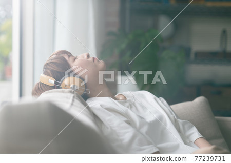 Young woman sitting on the sofa and listening to music on headphones 77239731