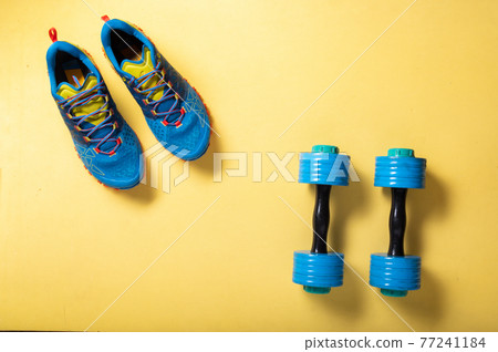 Sports blue shoes and blue dumbbells. Athletics at home. Sneakers and dumbbells on a yellow background. Top. Sports blue shoes and blue dumbbells. Athletics at home. Sneakers and dumbbells on a yellow background. Top. 77241184