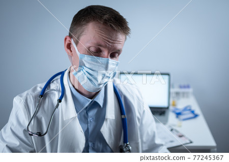 Tired young doctor takes off glasses and protective medical mask, looking at camera in hospital office on background of desktop. Topic is overburdened medical system at coronavirus, pandemic covid 19 77245367