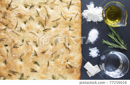 Recipe for cooking traditional Italian food - focaccia bread. The ingredients are placed to the right of the baked focaccia on a black board - flour, sugar, salt, yeast, olive oil, water, and rosemary 77256016