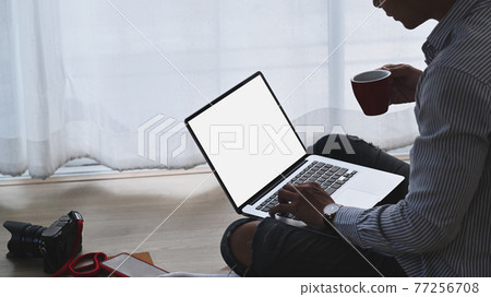 Cropped shot of freelance photographer is working with laptop while drinking coffee at his workplace. Cropped shot of freelance photographer is working with laptop while drinking coffee at his workplace. 77256708