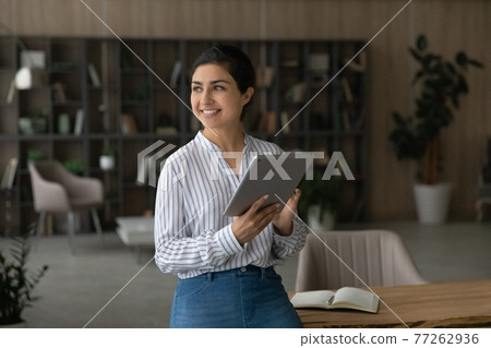 Dreamy smiling Indian businesswoman holding tablet, looking to aside 77262936